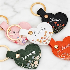 Puede incluir: Cinco llaveros en forma de corazón con anillas y broches de metal dorado. Los llaveros son negro, blanco, rosa, verde oscuro y naranja, cada uno con un estampado floral y un nombre en letra blanca o negra: Eleanor, Camila, Violet, Kennedy e Isabelle.
