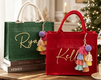 May include: Two rectangular tote bags made of textured fabric, one dark green and one red. Each bag features two cream-colored handles, gold cursive lettering, and a decorative keychain with a purple pom-pom and multi-colored fabric tassels.