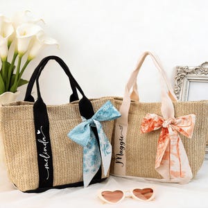 May include: Two woven tote bags with black and beige handles. One bag has a black band with the name "melinda" in script, and a blue patterned bow. The other bag has a beige band with the name "Maggie" and a peach-colored bow. Heart-shaped sunglasses are in front.