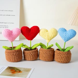 May include: Four crocheted heart-shaped flowers in brown pots. The hearts are pink, red, yellow, and blue. Each flower has green leaves and a green stem. The pots are a light brown colour.