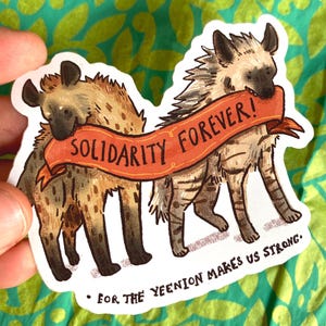 May include: A sticker featuring two cartoon hyenas with a red banner that reads "Solidarity Forever!" The banner is held by the hyenas' mouths. Below the hyenas, the text reads "For the Yeenion Makes Us Strong."