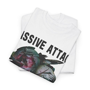 May include: White t-shirts with the text "ISSIVE ATTAC" in black, and a floral graphic with pink and green hues. The shirts are folded and stacked, showcasing the design on the top shirt.