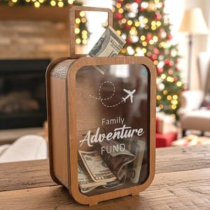 Op de afbeelding: Een houten koffer-vormige spaarpot met een doorzichtige voorkant, gevuld met contant geld. De spaarpot heeft een handvat en de tekst "Family Adventure Fund" met een vliegtuigafbeelding en stippellijn. De spaarpot staat op een houten tafel.