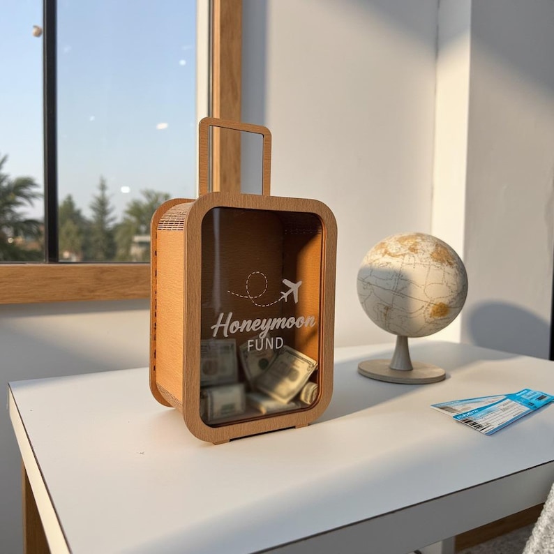 K&ouml;nnte beinhalten: Eine Geldbox in Form eines Holzkoffers mit einer Glasfront. Die Box hat einen Schlitz f&uuml;r Geld und tr&auml;gt die Aufschrift "Honeymoon Fund" auf der Vorderseite. Im Inneren der Box befinden sich Dollar-Scheine.