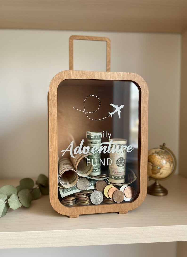 May include: Wooden travel-themed money box with a clear front. The box is filled with rolled-up bills and coins. White text on the front reads "Family Adventure Fund" with a graphic of an airplane and its flight path.