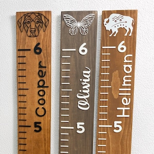 May include: Three wooden growth charts featuring geometric animal and insect cutouts. The first shows a dog and the name Cooper, the second a butterfly and Olivia, and the third a bison and Hellman. Each board displays height markings from 5 to 6.