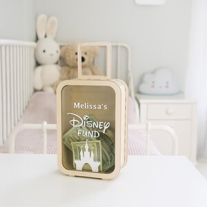 K&ouml;nnte beinhalten: Eine hellfarbene Geldkassette aus Holz in Form eines Koffers mit einem Griff oben. Die Box hat eine transparente Frontplatte und enth&auml;lt Papiergeld. Auf der Vorderseite sind die Worte "Melissa's Disney Fund" zusammen mit einer Schlosssilhouette aufgedruckt.