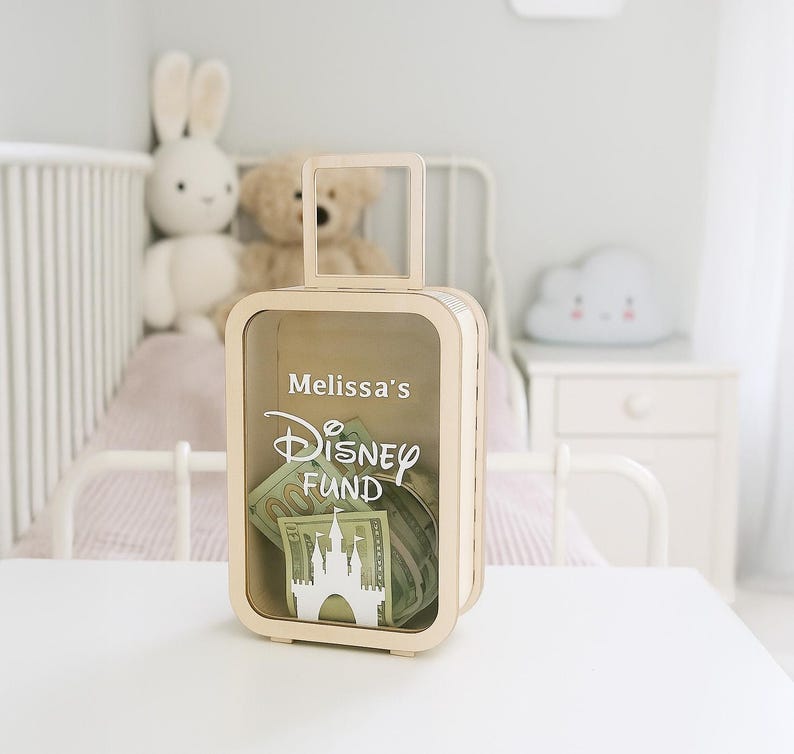 K&ouml;nnte beinhalten: Eine hellfarbene Geldkassette aus Holz in Form eines Koffers mit einem Griff oben. Die Box hat eine transparente Frontplatte und enth&auml;lt Papiergeld. Auf der Vorderseite sind die Worte "Melissa's Disney Fund" zusammen mit einer Schlosssilhouette aufgedruckt.