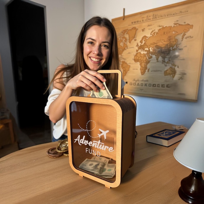 K&ouml;nnte beinhalten: Eine h&ouml;lzerne Sparb&uuml;chse in Form eines Koffers mit einer durchsichtigen Frontplatte. Die Box tr&auml;gt den Schriftzug "Adventure Fund" und ein kleines Flugzeug, das auf die Vorderseite gedruckt ist. Eine Person legt Geld in die Box.
