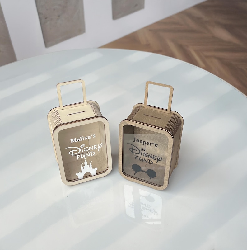 K&ouml;nnte beinhalten: Zwei Holzspardosen in Form von Koffern. Eine Box hat "Melisa's Disney Fund" und eine Burg, die andere "Jasper's Disney Fund" und einen Mickey Mouse Kopf. Beide haben einen Schlitz f&uuml;r Geld und einen Griff.