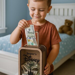 K&ouml;nnte beinhalten: Eine Holzspardose mit dem Text "Jasper's Disney Fund" und einer Schloss-Silhouette. Ein Kind steckt einen 100-Dollar-Schein in den Schlitz. Die Box ist mit verschiedenen W&auml;hrungen gef&uuml;llt.