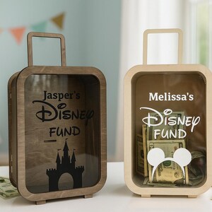 K&ouml;nnte beinhalten: Zwei Geldkassetten aus Holz in Form von Koffern. Eine ist braun und die andere hellbeige. Die braune Box hat "Jasper's Disney Fund" und eine Schlosssilhouette. Die beige Box hat "Melissa's Disney Fund" und einen Mickey Mouse-Kopf.