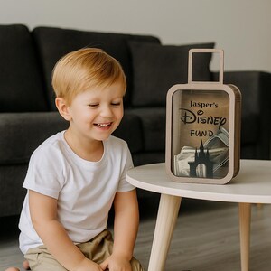 K&ouml;nnte beinhalten: Eine holzfarbene Spardose in Form eines Koffers mit dem Text "Jasper's Disney Fund" und einer Schloss-Silhouette. Ein Kind l&auml;chelt die Spardose auf einem wei&szlig;en Tisch an. Spielzeugautos und ein Dinosaurier liegen auf dem Boden.