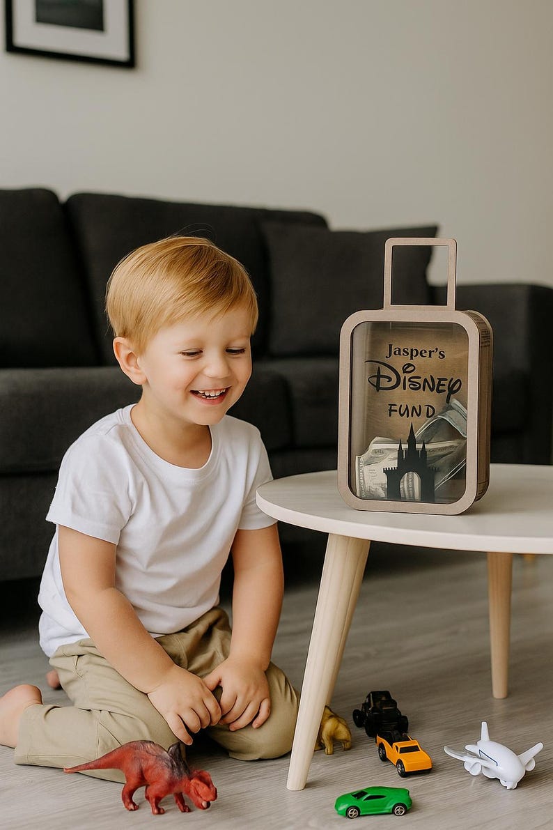 K&ouml;nnte beinhalten: Eine holzfarbene Spardose in Form eines Koffers mit dem Text "Jasper's Disney Fund" und einer Schloss-Silhouette. Ein Kind l&auml;chelt die Spardose auf einem wei&szlig;en Tisch an. Spielzeugautos und ein Dinosaurier liegen auf dem Boden.