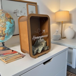 K&ouml;nnte beinhalten: Eine Geldbox in Form eines Holzkoffers mit einer Glasfront. Die Box hat einen Schlitz f&uuml;r Geld und tr&auml;gt die Aufschrift "Honeymoon Fund" auf der Vorderseite. Neben dem Text befindet sich ein kleines Flugzeugsymbol.