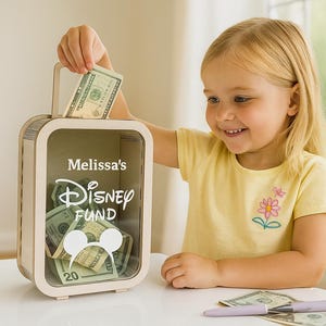 K&ouml;nnte beinhalten: Eine Holzspardose mit Griff, beschriftet mit "Melissa's Disney Fund", wird mit einem Zwanzig-Dollar-Schein gef&uuml;llt. Die Box ist rechteckig mit einer transparenten Vorderseite, und der Innenraum ist mit Bargeld gef&uuml;llt. Ein junges M&auml;dchen in einem gelben Hemd l&auml;chelt.