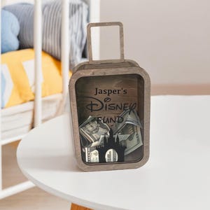 K&ouml;nnte beinhalten: Eine h&ouml;lzerne Spardose in Form eines Koffers mit Griff. Die Spardose tr&auml;gt den Text "Jasper's Disney Fund" und eine Schloss-Silhouette. Die Spardose ist mit Banknoten gef&uuml;llt.