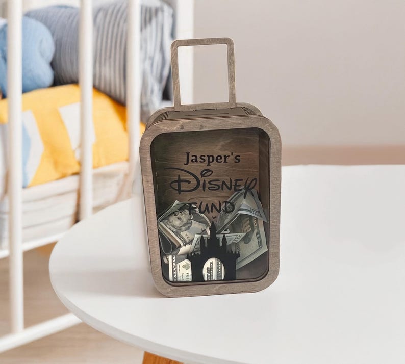 K&ouml;nnte beinhalten: Eine h&ouml;lzerne Spardose in Form eines Koffers mit Griff. Die Spardose tr&auml;gt den Text "Jasper's Disney Fund" und eine Schloss-Silhouette. Die Spardose ist mit Banknoten gef&uuml;llt.