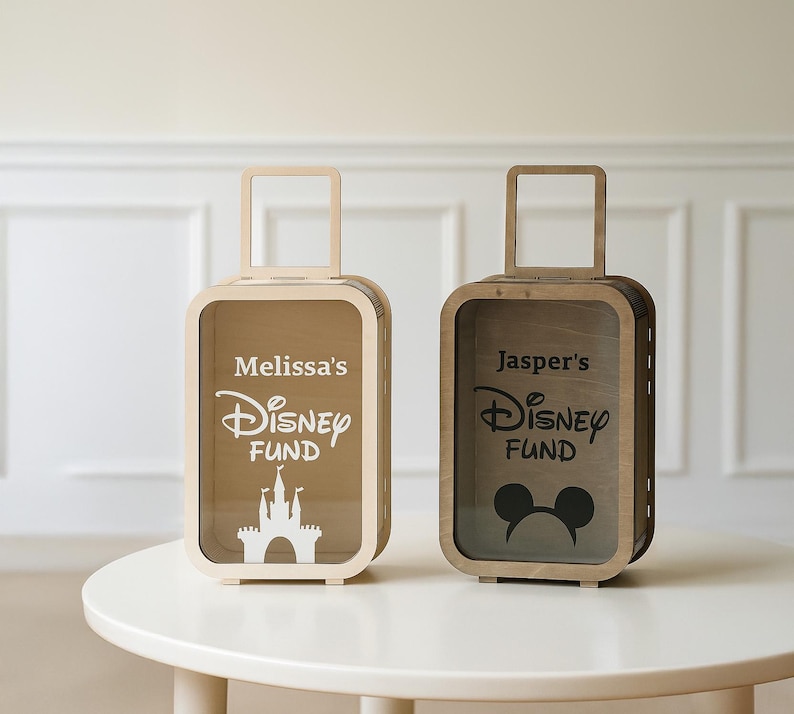 K&ouml;nnte beinhalten: Zwei holzfarbene Spardosen in Form von Koffern mit transparenten Fronten. Eine Box tr&auml;gt die Aufschrift "Melissa's Disney Fund" mit einem Schloss, die andere "Jasper's Disney Fund" mit Mickey Mouse Ohren. Beide haben Griffe.