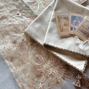以下が含まれることがあります： 金色の刺繍とスパンコールが施された白いレースのテーブルクロス。白いベルベットの布が、金色のトリムとタッセルで飾られ、テーブルクロスの上に置かれています。ベルベットの布の上にタロットカードが3枚置かれています。