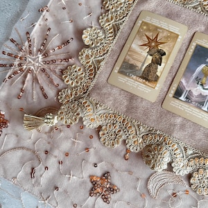 以下が含まれることがあります： 星と月のデザインが施された、ピンクとゴールドのタロットクロス。ゴールドレースのトリムが施されています。クロスの上には、「星」と「カップのエース」の2枚のタロットカードが置かれています。