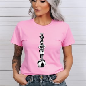 Puede incluir: Una camiseta rosa con una ilustración en blanco y negro de una mujer haciendo equilibrio sobre una pelota. La mujer lleva un sombrero y tiene un mono en la cabeza que sostiene un paraguas.