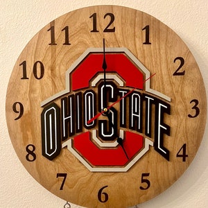 May include: A wooden clock with a red, white, and gray Ohio State logo. The clock face has black numbers 1 through 12.