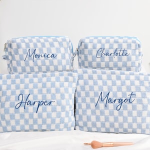 May include: Four light blue and white checkered cosmetic bags with names embroidered in a cursive font: Monica, Charlotte, Harper, and Margot. The bags are zippered and sit on a white surface.