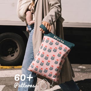 May include: A rectangular zippered pouch with a pink background featuring a repeating pattern of dark green stems and coral-colored tulip-like flowers. The top section is solid dark green with the name Maria printed in white. It has a wrist strap.