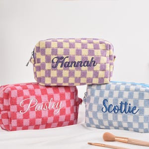 Puede incluir: Tres neceseres a cuadros en morado, rojo y azul. Cada bolsa tiene un nombre bordado: "Hannah", "Paisley" y "Scottie". Hay brochas de maquillaje delante de las bolsas.