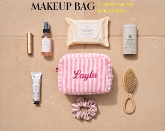 May include: A pink and white striped plush makeup bag with the name Layla embroidered in pink script. The bag is surrounded by various beauty items including a spray bottle, a tube, a pack of wipes, a deodorant stick, a hairbrush, and a scrunchie.