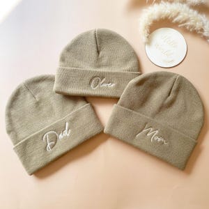 Puede incluir: Tres gorros de punto beige con texto bordado en blanco. Los gorros dicen "Papá", "Mamá" y "Oliver".