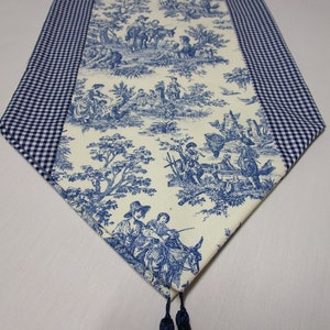 May include: A blue and white table runner with a toile pattern and gingham trim. The runner has a tassel at the end.