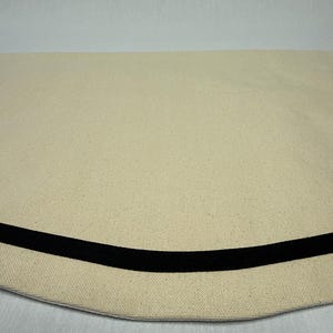 May include: A semi-circular piece of off-white canvas fabric featuring a smooth, curved edge. A single, solid black ribbon or trim is stitched horizontally across the lower section of the fabric, creating a distinct contrast against the light material.