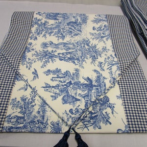 May include: A blue and white toile table runner with a gingham border and tassels. The runner features a French country design with a white background and blue floral patterns.