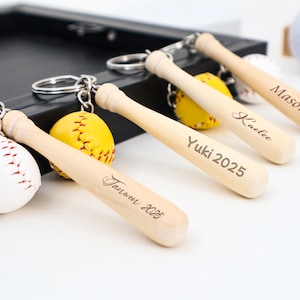 May include: Baseball-themed keyrings with miniature wooden bats and baseballs. The bats are engraved with names and the year 2025. One keyring features a white baseball with red stitching, others have yellow baseballs.