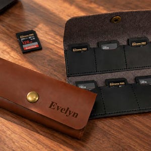 May include: A brown leather memory card holder with a brass snap closure and the name Evelyn engraved on the front flap. The interior is black with six individual slots, each containing a memory card, with one additional card lying nearby.
