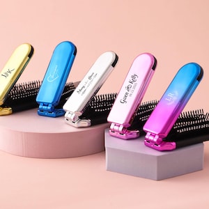 May include: A selection of personalised folding hairbrushes in gold, blue, white, pink, and a pink-to-blue gradient. Each brush has a different inscription, including initials and names with dates. The brushes are displayed on pink and grey platforms.