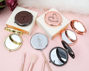 May include: A collection of personalized compact mirrors in various colors, including gold, rose gold, silver, and black. Each mirror features a unique design, such as an initial, a heart, or a lightbulb with floral elements. Makeup brushes are also visible.