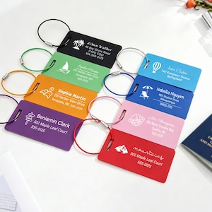Pode incluir: Um conjunto de oito etiquetas de bagagem retangulares nas cores preta, verde, laranja, roxa, azul, azul-claro, rosa e vermelha. Cada etiqueta apresenta um ícone, nome, endereço e número de telefone, fixada por um aro de metal circular.