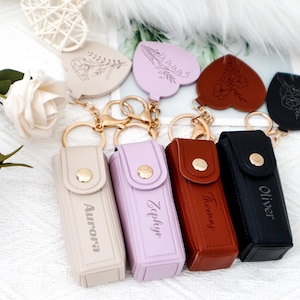 Può includere: Una collezione di portachiavi personalizzati con custodie rettangolari e ciondoli a forma di cuore. Le custodie sono beige, lavanda, marrone e nere, ognuna incisa con un nome. I ciondoli a cuore hanno disegni floreali. Hardware color oro.