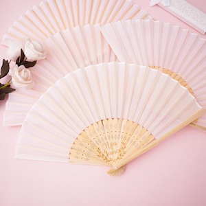 以下が含まれることがあります： ピンク色の表面に、木製の柄が付いた、開いた薄いピンク色の紙扇子がいくつか配置されています。 扇子の1つには、金色の文字で「Ava James」と書かれています。白いバラと小さなギフトボックスも表示されています。