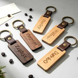 May include: Five rectangular wooden keychains with dark brown leather straps and brass-toned metal rings. Each keychain is engraved with a name: Zephyr, Caspian, Harley, Willow, and Dumbo. The wood tones vary from light to dark brown.