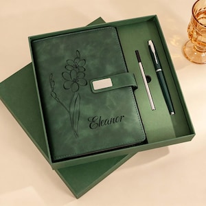 May include: A dark green notebook with a floral line art design and the name Eleanor engraved on the cover. The notebook features a magnetic closure with a silver accent and is presented in a matching green gift box with a pen and a refill.
