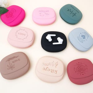 May include: A selection of colourful, pebble-shaped cases for wireless earbuds. The cases are in various colours, including pink, black, and teal, and feature embossed designs and text. One case is open, displaying white earbuds.