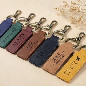 May include: A collection of personalized leather keychains in various colors, including blue, green, burgundy, brown, navy, and yellow. Each keychain features engraved text and designs, such as names, initials, and dates. The keychains have silver-tone metal hardware.