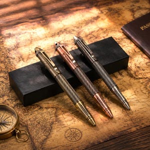 May include: Three metal pens with a twisted, textured grip and a smooth, pointed tip. Each pen features a clip and a personalized name engraved on the barrel. The pens are finished in brass, copper, and gunmetal tones, resting on a black rectangular box.