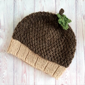 May include: A brown and beige knitted hat with a green leaf detail on top. The hat is made of a soft, textured yarn and has a simple, classic design.