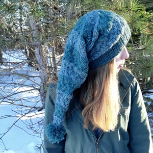 Puede incluir: Un gorro de punto azul y gris con una larga parte superior puntiaguda y un pompón. El gorro lo lleva puesto una persona con el pelo largo y castaño.