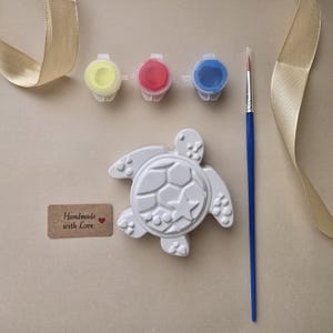 May include: A white turtle-shaped plaster figure with a geometric shell pattern, three small paint pots in yellow, red, and blue, a thin blue paintbrush, and a rectangular brown tag with the text Handmade with Love and a small red heart.